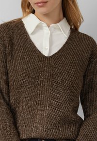 Kobieta w brązowym swetrze z prążkowanej dzianiny w serek, założonym na białą koszulę z kołnierzykiem, z luźnymi blond włosami.