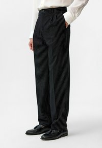 Zwarte pinstriped broeken met een hoge taille, plooien en zijzakken, gemaakt van soepel stof; gecombineerd met zwarte loafers en een wit shirt.