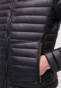 Svart pufferjacka med quiltat mönster, dragkedja framtill och fickor, slät textur och lätt material.