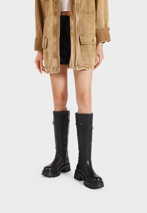 Stivali in pelle nera al ginocchio con una suola spessa e dettagli in coulisse discreti, abbinati a un giubbotto beige e pantaloni corti neri.