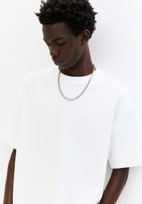 Hvid oversized T-shirt med korte ærmer, lavet af blødt stof. Har en rund halsudskæring og et sølv kædehalssmykke som accent.