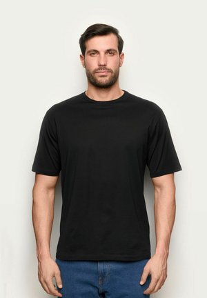 Uomo in piedi davanti a uno sfondo uniforme, indossa una maglietta nera a maniche corte semplice e jeans blu, guarda avanti con un'espressione neutra.