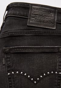 Jean en denim noir Levi’s en gros plan montrant la poche arrière avec coutures cloutées et un patch en cuir de la marque au-dessus de la taille.