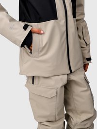Persoană cu mâna în buzunar, purtând o geacă de exterior bej și neagră și pantaloni cargo bej, pe un fundal simplu.