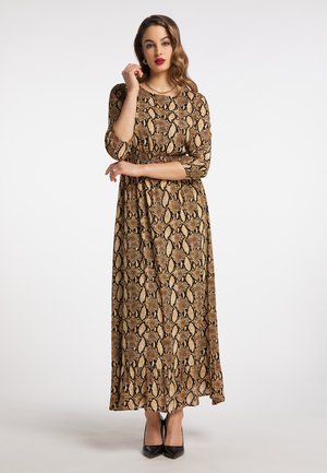 Donna che indossa un vestito marrone a maniche lunghe e lungo fino ai piedi con stampa a motivo serpente e tacchi neri, posando con una mano vicino al viso e l'altra incrociata.