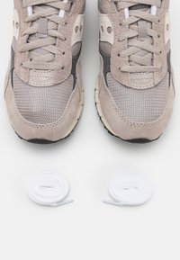 Sneaker in suede beige e mesh con suole nere, caratterizzati da un design con lacci. Due lacci bianchi sono posizionati sotto le scarpe.