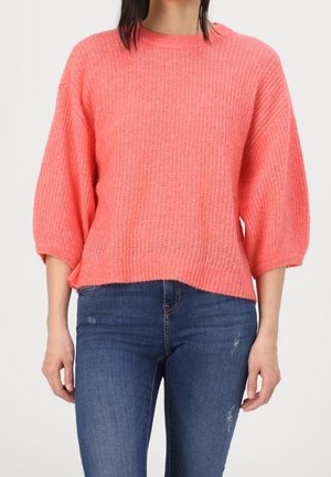 Femme portant un pull ample en maille corail rose avec des manches 3/4, associé à un jean skinny bleu déchiré.