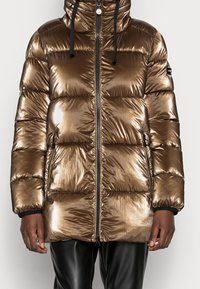 Chaqueta acolchada metálica bronce con capucha, cuerdas negras, cremallera frontal, bolsillos con cremallera, llevada por una persona de piel oscura y pantalones negros.