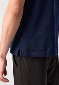 Camisa de manga curta azul-marinho com um padrão texturizado, fendas laterais e um pequeno rótulo da marca na orla. Usada com calças escuras.