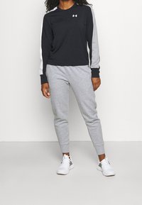 Maglietta nera a maniche lunghe con dettagli grigi e logo, abbinata a pantaloni jogger grigi con orlo a coste e scarpe da ginnastica bianche.