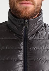 Graue Pufferjacke mit hohem Kragen, vertikal gestepptem Design, Reißverschluss und glatter, glänzender Textur. Mit einem dunklen Reißverschluss.