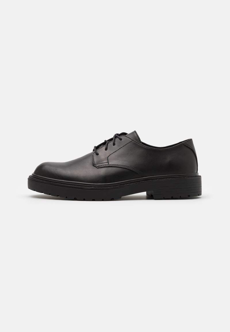 Chaussure habillée pour homme en cuir noir à lacets, avec semelle épaisse en caoutchouc et petit talon, présentée de profil sur un fond clair uni.