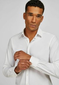 Chemise blanche avec col et poignets boutonnés. Tissu tissé lisse avec une coupe ajustée. Présente des accents de boutons sur les poignets.