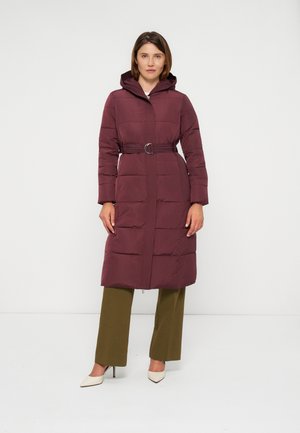 Femme portant un long manteau matelassé bordeaux ceinturé avec une capuche, un pantalon ample vert olive et des talons blancs pointus, debout devant un fond blanc.