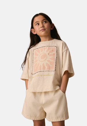 Girl with long dark hair wearing beige shorts and a loose beige t-shirt featuring a sun graphic and "sea full of sunshine" text.