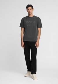 Camiseta gris de cuello redondo con el texto grabado "POLO CLUB", combinada con pantalones negros y zapatillas beige, sobre un fondo claro y liso.
