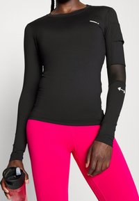 Schwarzes langärmliges Workout-Oberteil mit enganliegendem Design und durchsichtigen Ärmeldetails, kombiniert mit leuchtend pinkfarbenen Leggings. Inklusive einer Wasserflasche.