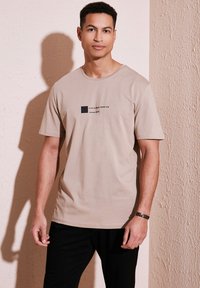 Šviesiai ruda medvilninė t-shirt su trumpomis rankovėmis, su juodu grafiniu tekstu "VISADA ŠYPSOKIS NEEKITE LIŪDNAS" ir mažu juodu kvadratiniu piešiniu, atspausdintu ant krūtinės.