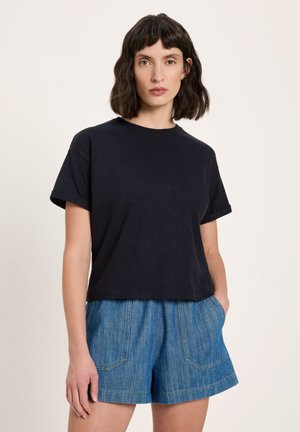 Woman with shoulder-length dark hair wears black short-sleeve T-shirt and high-waisted blue denim shorts, standing with one hand in pocket.