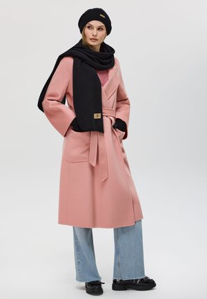 Cappotto in misto lana rosa con maniche larghe, cintura e tasche; abbinato a una sciarpa nera e un cappello lavorato a maglia; indossato con pantaloni flared in denim chiaro e stivali neri.