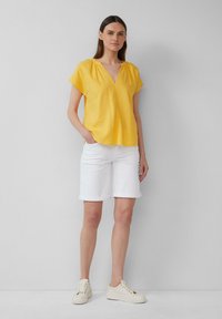 Haut en lin jaune à manches courtes et col en V, associé à un short blanc à revers. Le mannequin porte des baskets beiges. Fond uni.