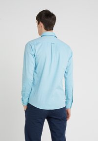 Jasnoniebieska koszula z długim rękawem z miękkiego materiału, z klasycznym kołnierzykiem, zapięciem na guziki na mankietach i zakrzywionym dołem, pokazana od tyłu.