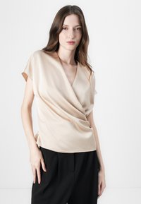 Beige, kortermet innpakningsgenser i glatt stoff med V-hals og drapert frontdetalj, kombinert med svarte bukser.