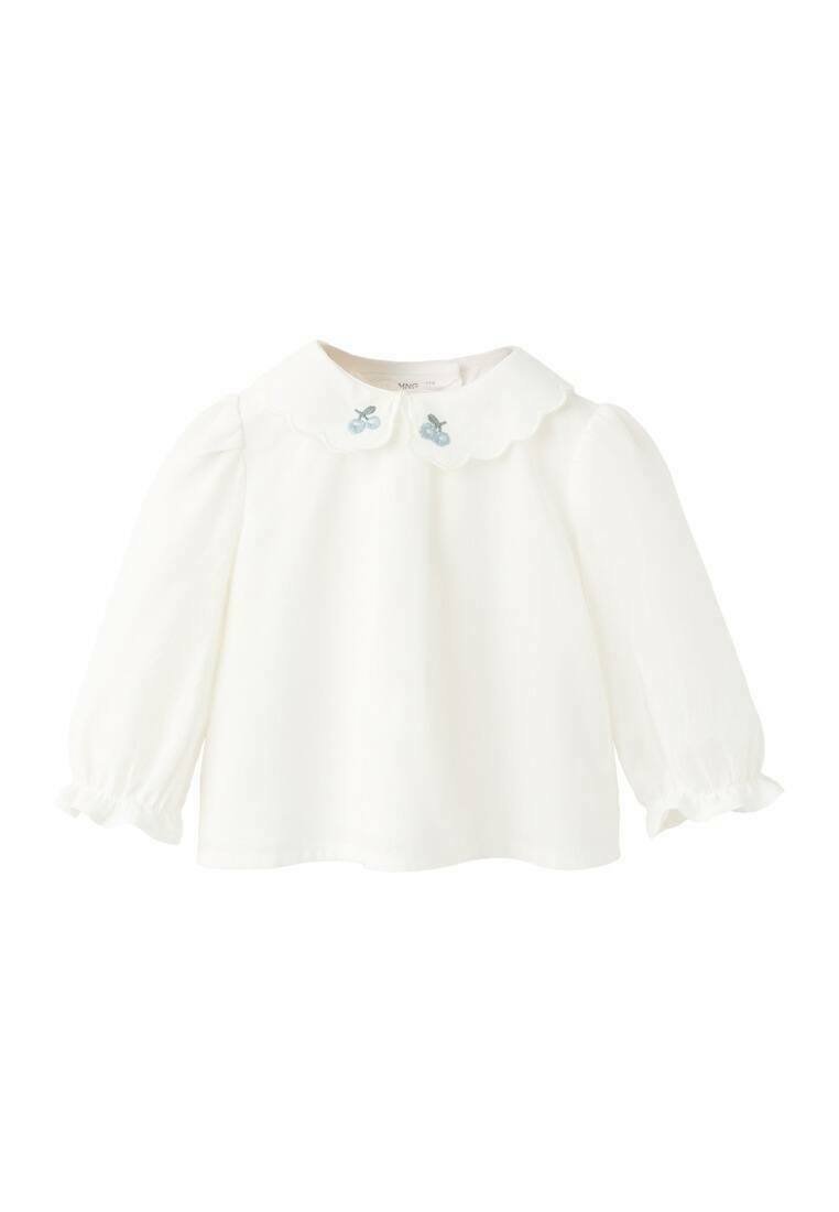 Bela majica z dolgimi rokavi z nazobčanim ovratnikom in svetlo modrimi vezenimi oblikami češenj. Mehka tkanina z nabranimi rokavi pri manšetah.