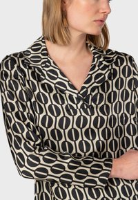 Blusa in satin con un motivo geometrico nero e beige, caratterizzata da un colletto a risvolto e maniche lunghe. Texture morbida e vestibilità rilassata.