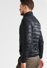 Jachetă puffer neagră cu guler cu fermoar, având un design matlasat și o textură netedă. Ușoară, cu o potrivire strânsă pe spatele purtătorului.