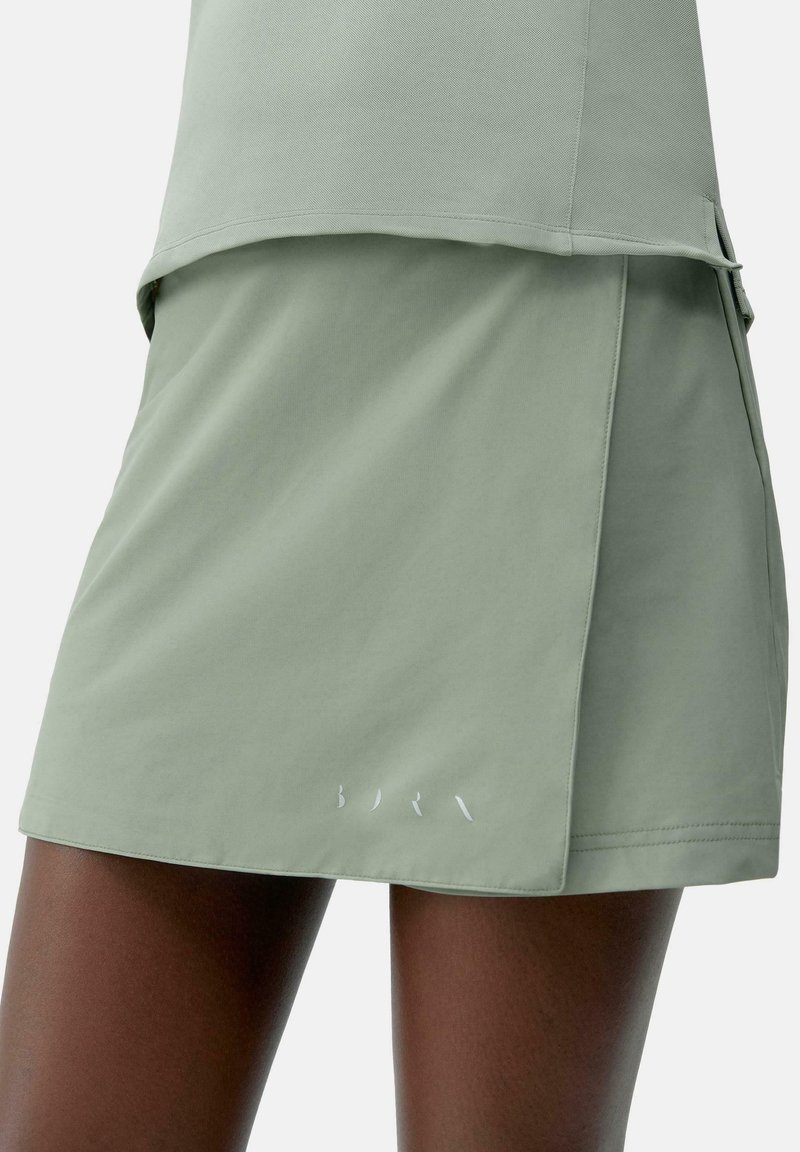 Een lichtgroene rok van zachte stof, met een gelaagd ontwerp en een klein wit logo aan de zoom. Soepele textuur, knielange snit.