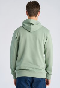 Man draagt een lichtgroene hoodie en blauwe spijkerbroek, staat met de rug naar de camera tegen een effen lichtgrijze achtergrond.