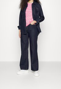 Žena v tmavě modré džínové bundě a širokých kalhotách, s tričkem s růžovými a bílými pruhy a bílými teniskami, pózující s jednou rukou v kapse.