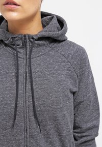 Primer plano de una persona que lleva una sudadera con cremallera de color gris oscuro y cordones, mostrando la parte inferior de la cara y la parte superior del torso sobre un fondo blanco.