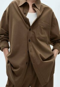 Chemise oversize marron à rayures fines avec un devant à boutons, des manches longues retroussées aux poignets et une poche poitrine ; associée à un débardeur blanc.