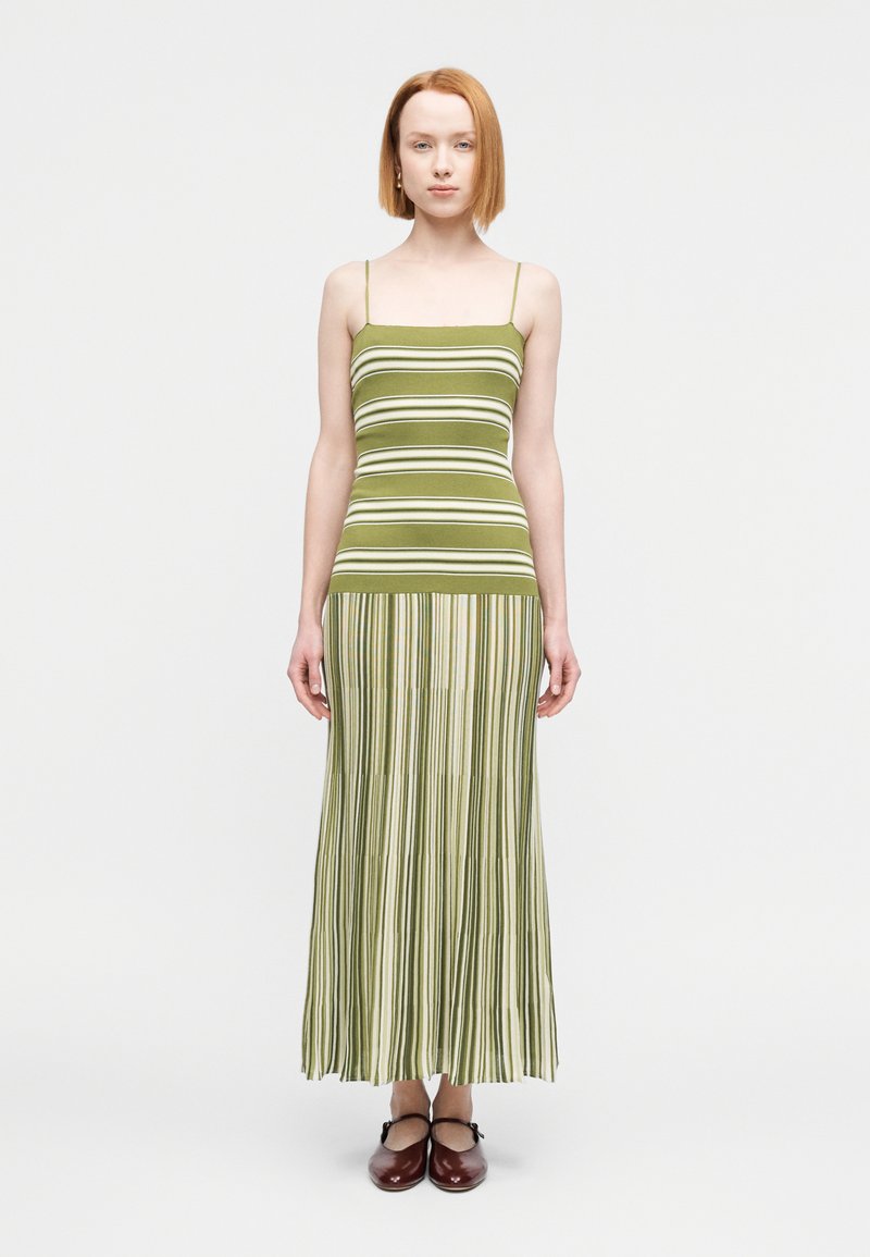 Νεαρή γυναίκα με ανοιχτόχρωμο δέρμα και κοντά κόκκινα μαλλιά, που φοράει μάξι φόρεμα με πράσινες και λευκές ρίγες χωρίς μανίκια και καφέ παπούτσια τύπου Mary Jane.