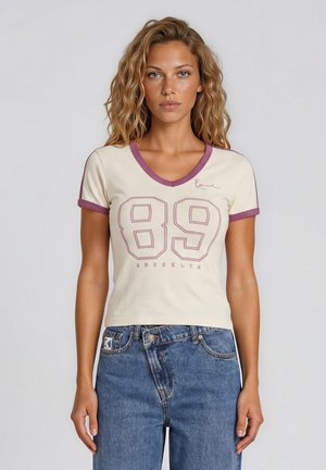 Femme aux cheveux ondulés portant un haut crème à manches courtes en V avec bordure violette, numéro 89 et texte « Brooklyn », associé à un jean bleu.