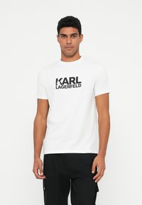 Valge puuvillane T-särk, mille esiosas on suur must "KARL LAGERFELD" logo, ümmargune kaelus ja lühikesed varrukad.