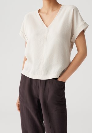 Frau trägt eine lockere, kurzärmelige cremefarbene V-Ausschnitt-Bluse mit umgeschlagenen Ärmeln, in hoch taillierte dunkelbraune Hosen gesteckt, Hände in den Taschen.