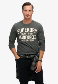 Grijze lange mouwen shirt met "Superdry Heavy Duty Reinforced Denim Comp" in crème gedrukt. Zwarte skinny jeans. Geruit overhemd om de taille geknoopt.