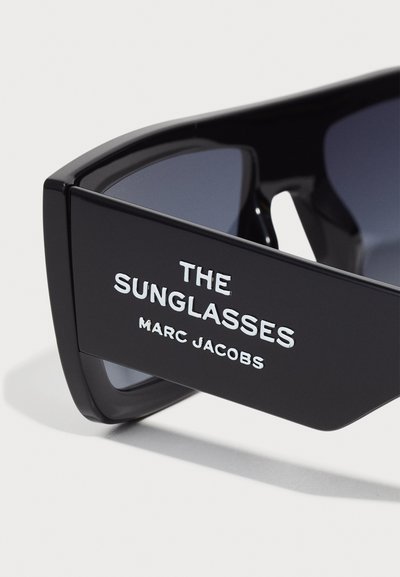 Schwarze Sonnenbrille mit eckigem Design, dunklen Gläsern und fettem weißen Text, der "DIE SONNENBRILLE" und "MARC JACOBS" an den Bügeln zeigt.