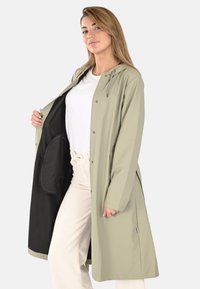 Manteau léger long couleur khaki avec capuche, boutons pression et fentes latérales. Doublure intérieure en tissu noir contrastant. Porté sur un haut blanc.