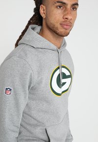 Sudadera con capucha gris, con un gran logo verde y amarillo en la parte frontal, cuenta con la marca de la NFL en la manga izquierda y capucha con cordón.