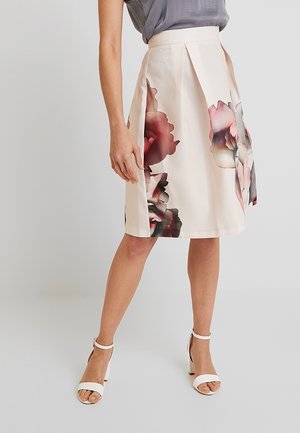 Mujer con falda crema hasta la rodilla con grandes estampados florales desvaídos y sandalias blancas de tacón alto, de pie sobre un fondo liso.