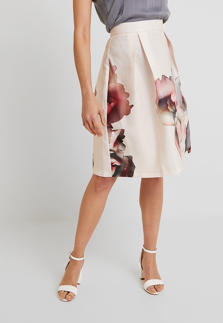 Femme portant une jupe crème longueur genou avec de grandes impressions florales délavées et des sandales blanches à talons hauts, debout sur un fond uni.