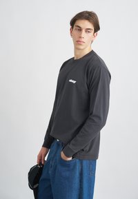 T-shirt de manga longa cinza escuro com decote redondo e logotipo branco. Combinado com jeans azuis largos, com bolsos laterais.