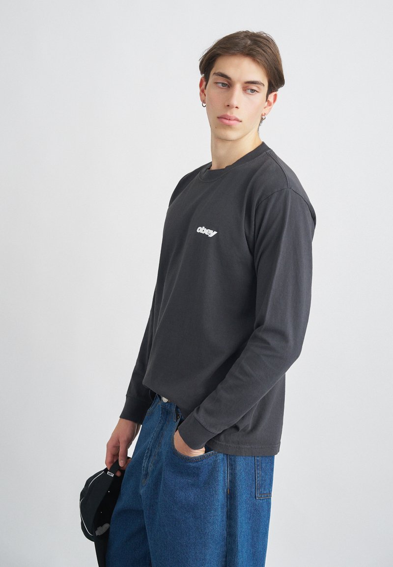 T-shirt de manga longa cinza escuro com decote redondo e logotipo branco. Combinado com jeans azuis largos, com bolsos laterais.