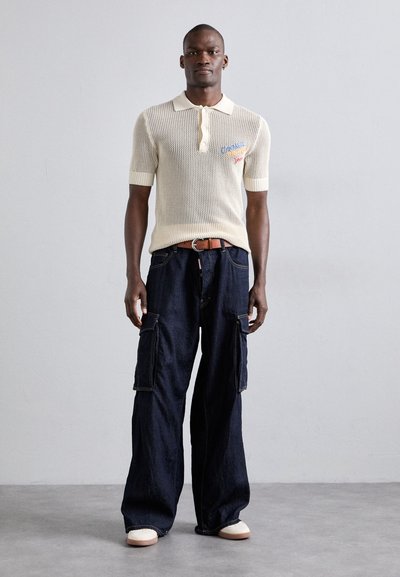 Short-sleeve beige knitted polo with three buttons and embroidered accents, paired with loose-fitting dark blue cargo pants featuring pockets.