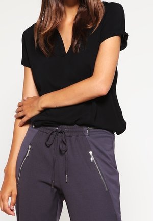 Femme portant un haut noir à manches courtes et un pantalon gris foncé à cordon avec poches zippées, posant avec un bras croisé sur le corps.