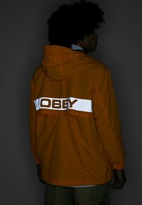 Orange hooded jacket featuring a white reflective stripe with the word "OBEY." Lightweight, with a casual fit and visible stitching details.